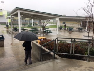 A campus supervisor is seeking shelter under the overhang, while one student is walking to their 6th period class in the pouring rain.