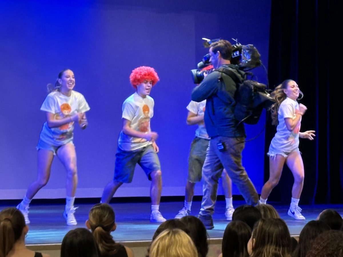 Student dancers performing on stage as the KTLA camera crew film for their live morning news show. The audience is filled with live spirited students cheering them on.